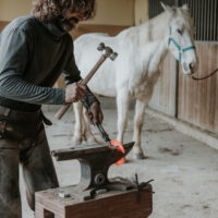 A farrier shapes a heated horse shoe for the horse in the background.