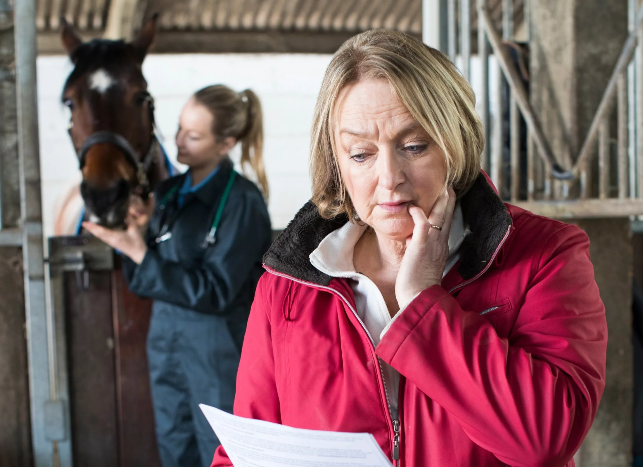 A woman in a red coat looks at a piece of paper with concern, <a href=