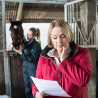 A woman in a red coat looks at a piece of paper with concern, while a horse is in the background.