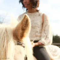 A woman wearing a cream colored sweater sits on a horse.