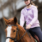Girl in a lavender hooded shirt and helmet while riding her brown horse in a riding lesson.